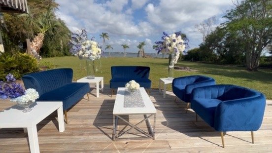 Navy and White Outdoor Lounge Setup with Coastal Backdrop Navy velvet lounge furniture with white tables and floral accents on a wooden deck overlooking palm trees and ocean.