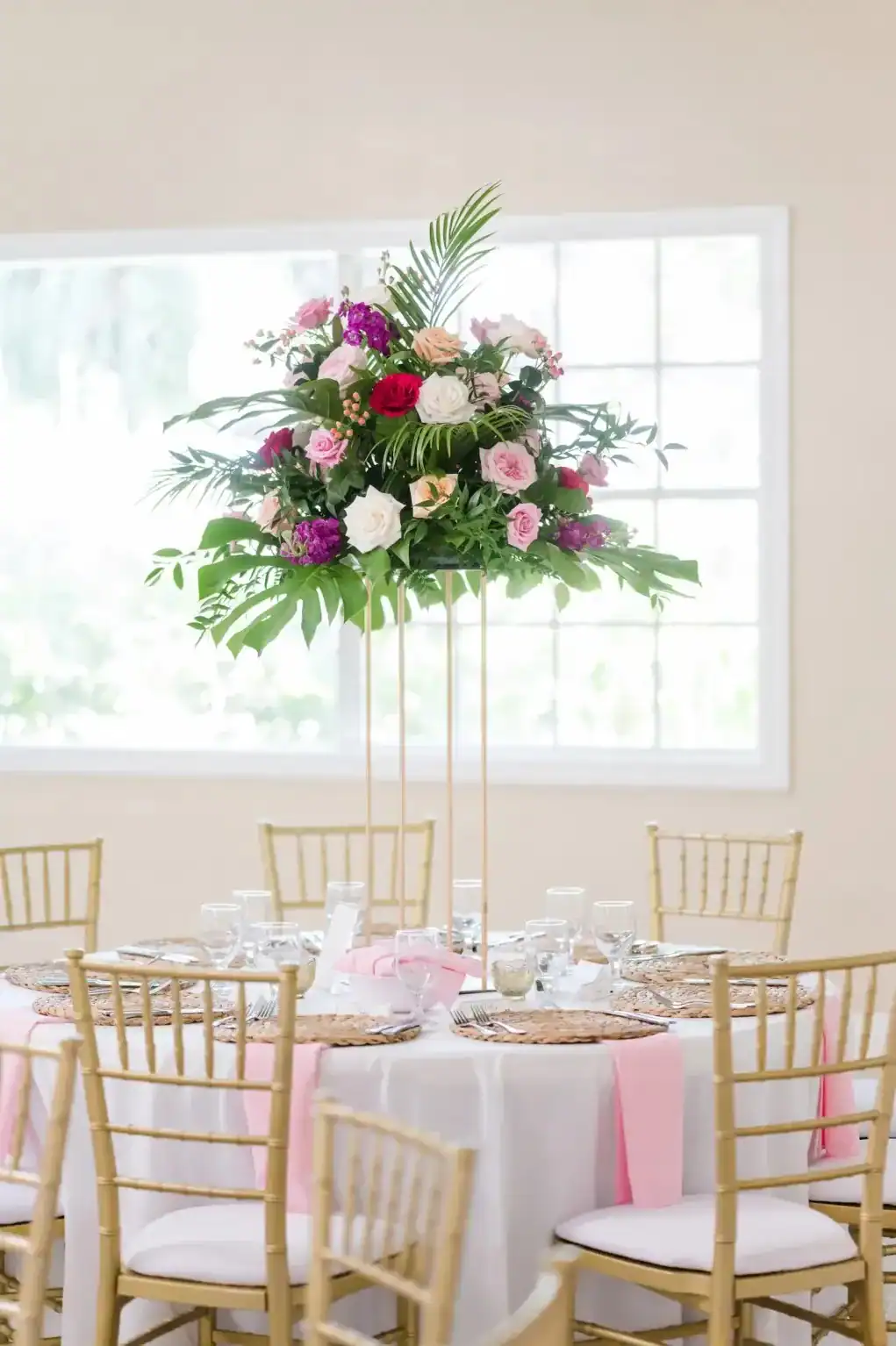 Tall pink, orange, white, and purple floral centerpiece above a reception table with gold chairs