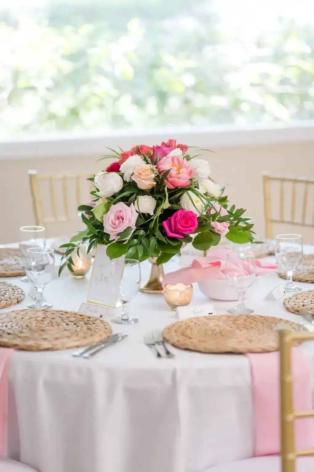 Round reception table with soft pink linens, woven chargers, colorful flowers, and gold chairs