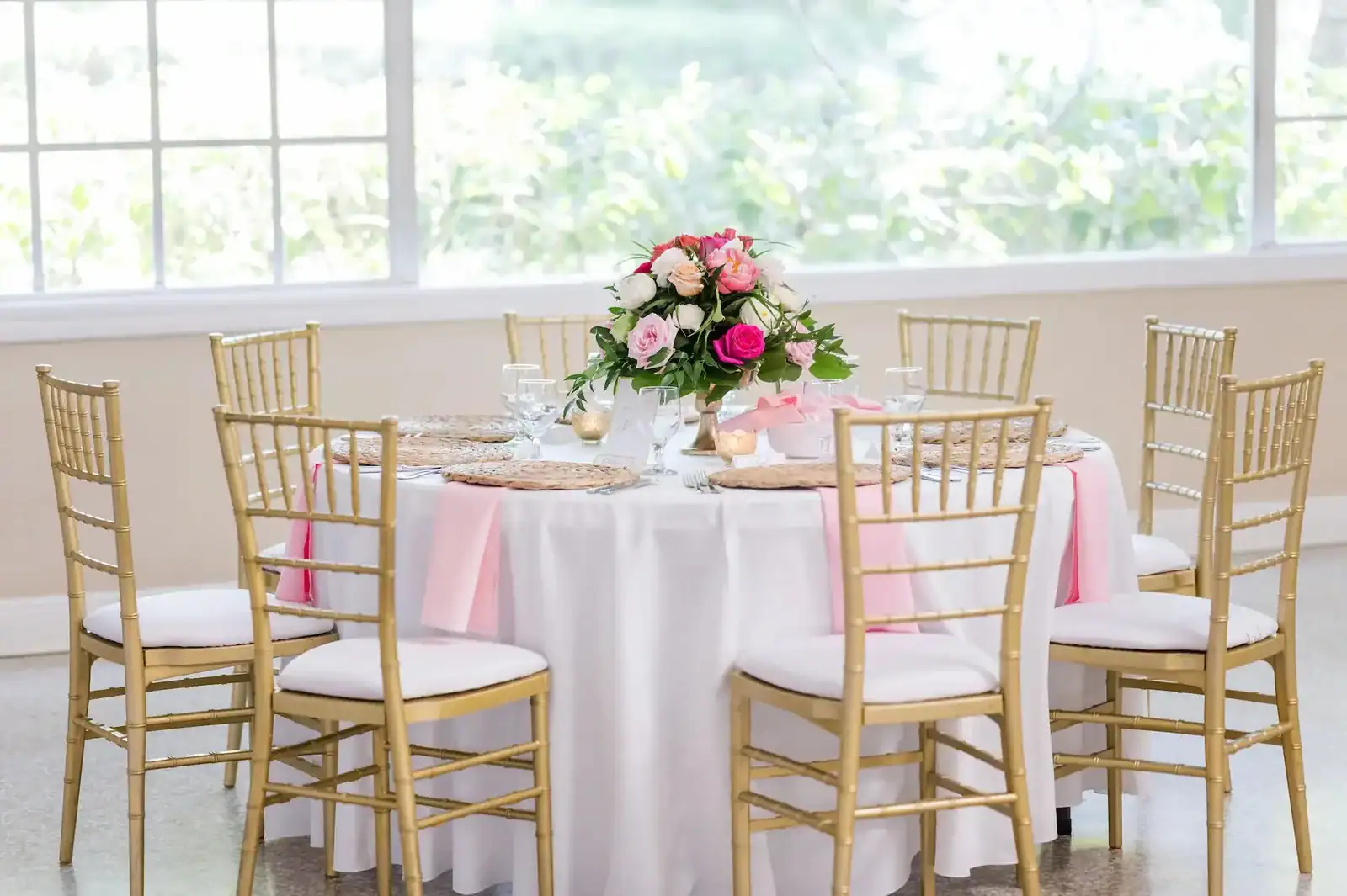 Vibrant waterfront reception table styled with pink linens, colorful flowers, woven chargers, and gold chairs
