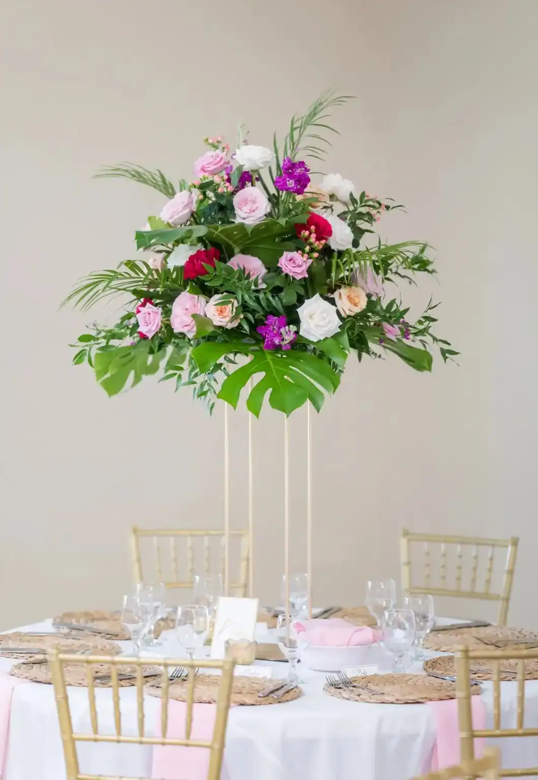 Tall tropical floral centerpiece above a round reception table with pink accents and gold chairs