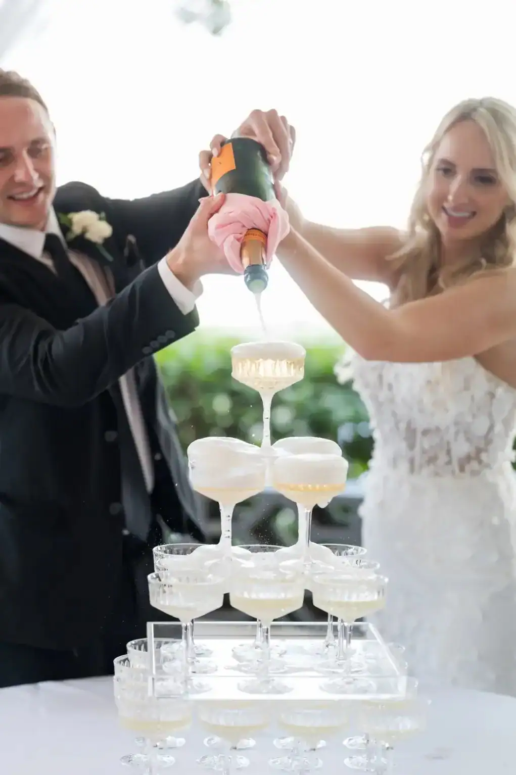 Katie and Alex pour champagne into a tiered glass tower during their colorful waterfront wedding celebration