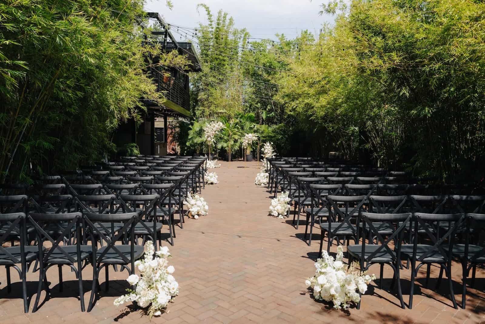 Rows of black chairs line a brick courtyard aisle with white floral arrangements at NOVA 535.