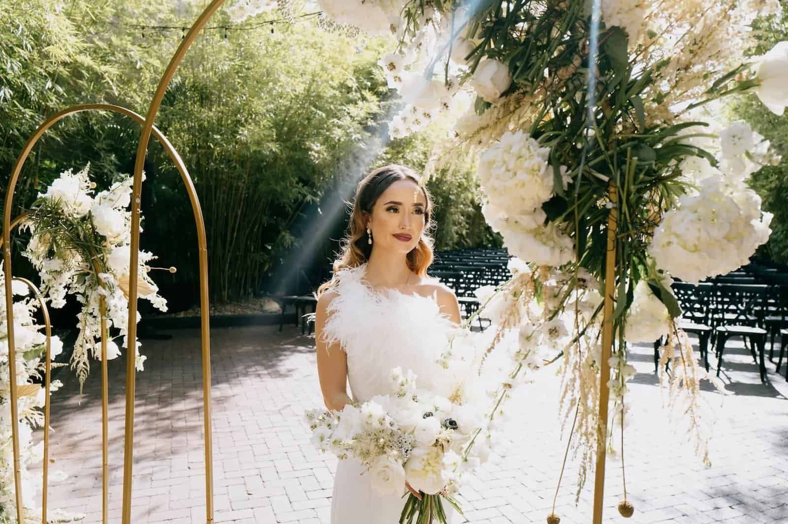 A bride stands before a gold ceremony arch decorated with white floral arrangements at NOVA 535.