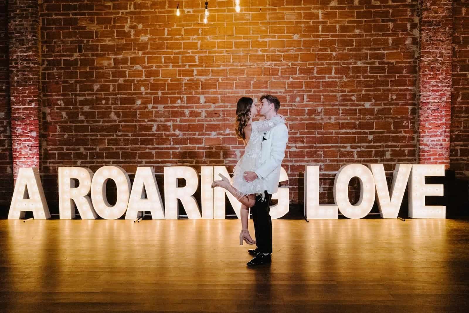 A couple kisses before an exposed brick wall and illuminated marquee letters spelling A Roaring Love.