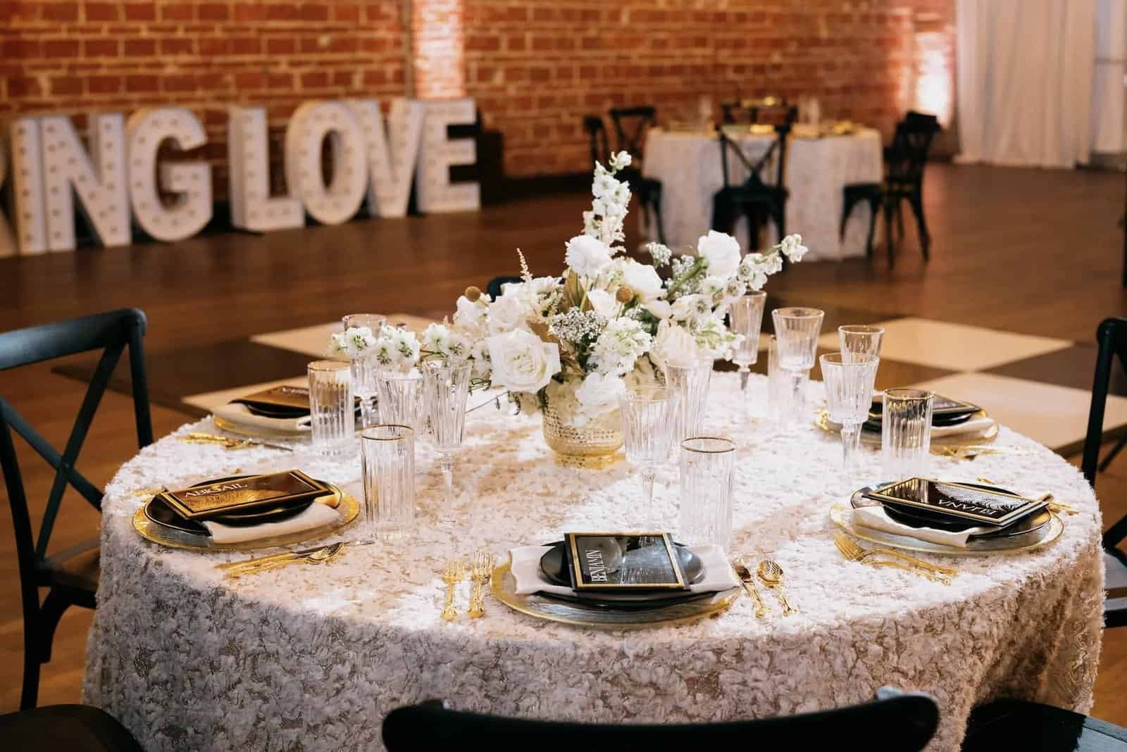 A round table is set with textured white linen, black plates, gold flatware, and white flowers.