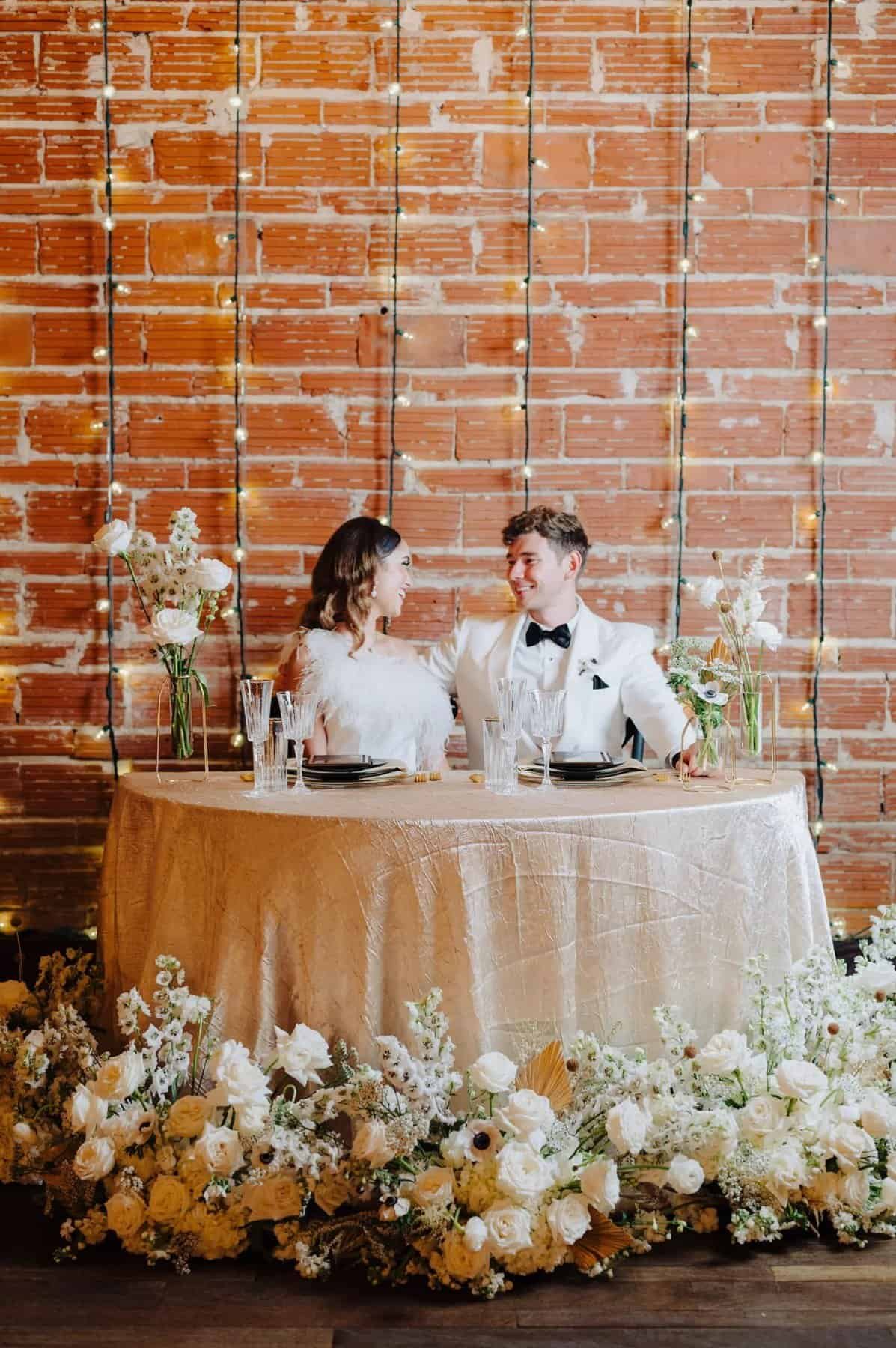 A couple sits at a sweetheart table with gold textured linen and white floral arrangements against a brick wall.