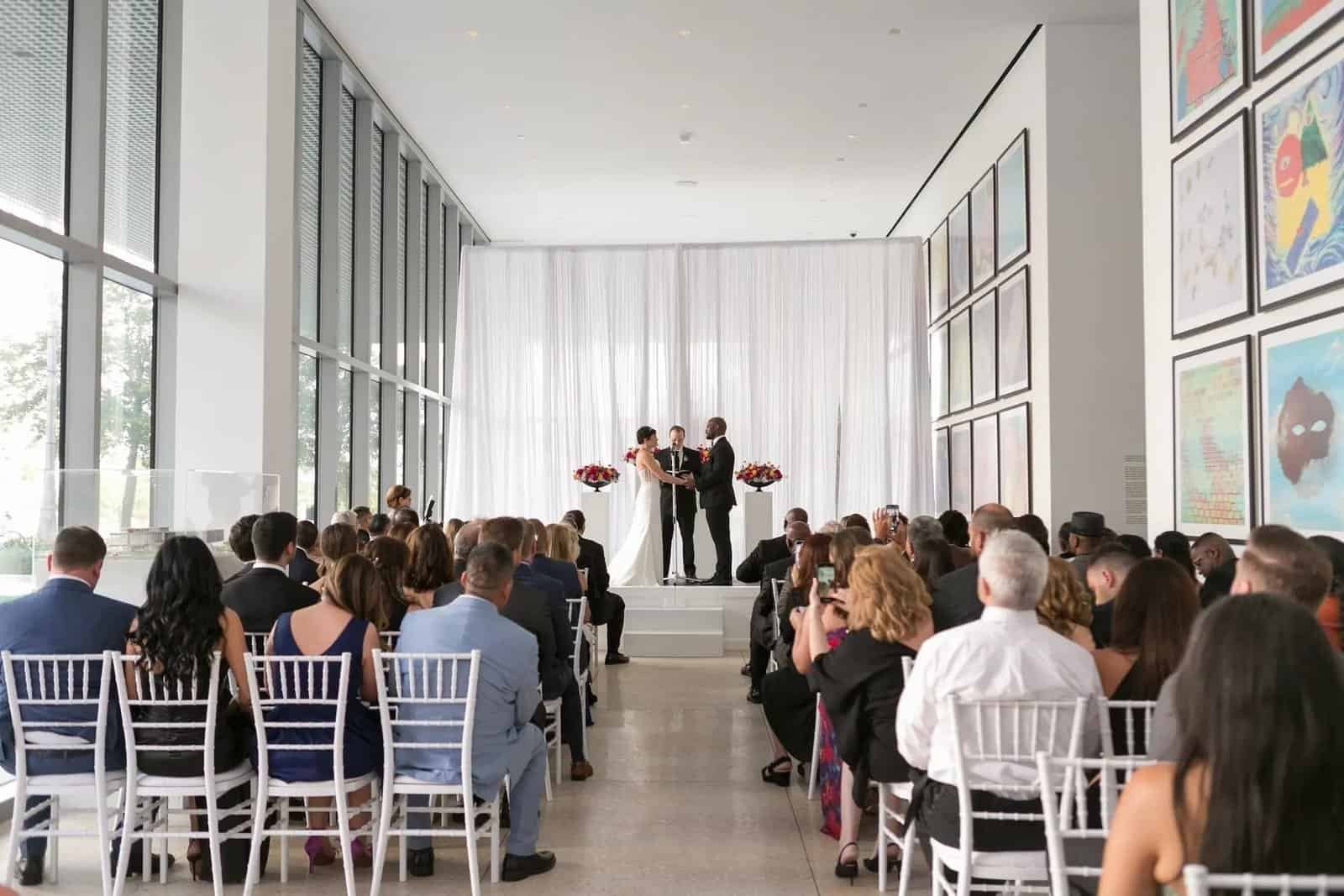 Guests seated on white chiavari chairs during the ceremony in the museum's gallery corridor