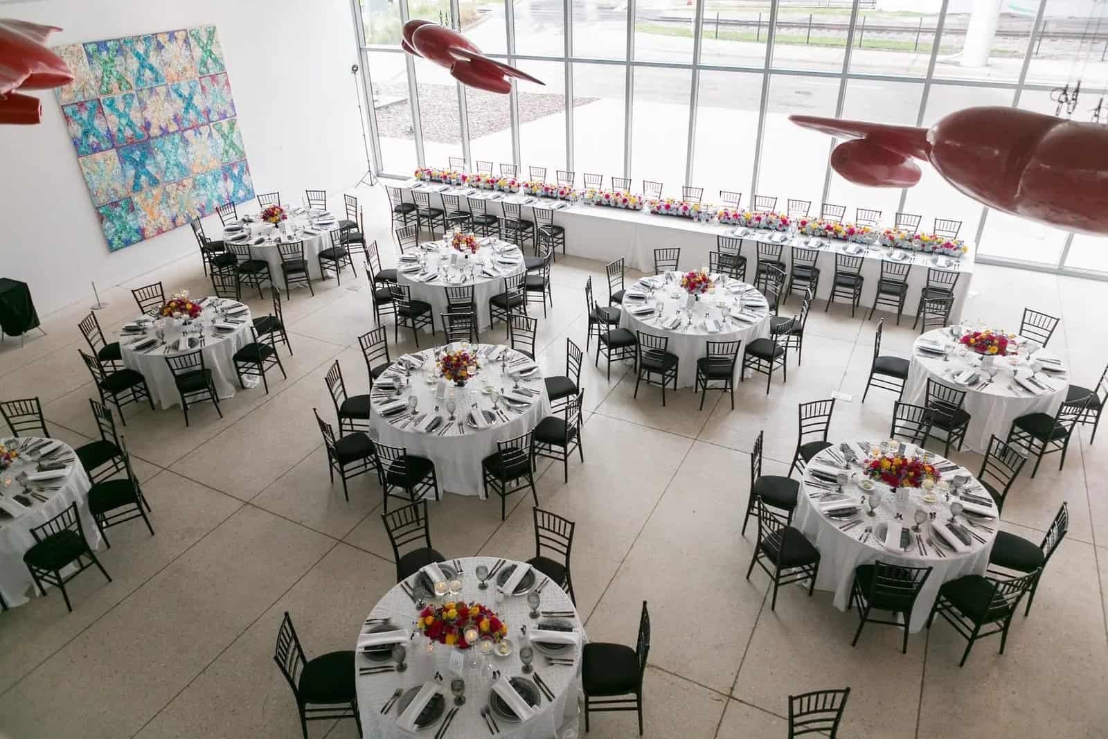 Reception round tables with white linens, black chiavari chairs and bright low centerpieces beneath the museum's red ceiling sculpture