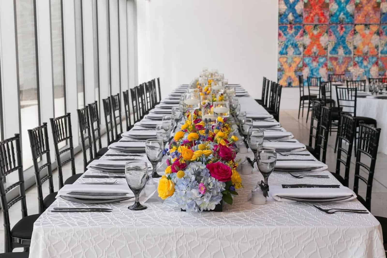 Long feasting table with black chiavari chairs and a runner of yellow, pink and blue blooms