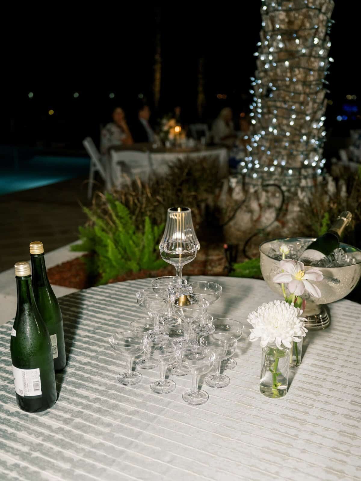 Acrylic champagne coupes and a bottle on a poolside table at an evening wedding reception
