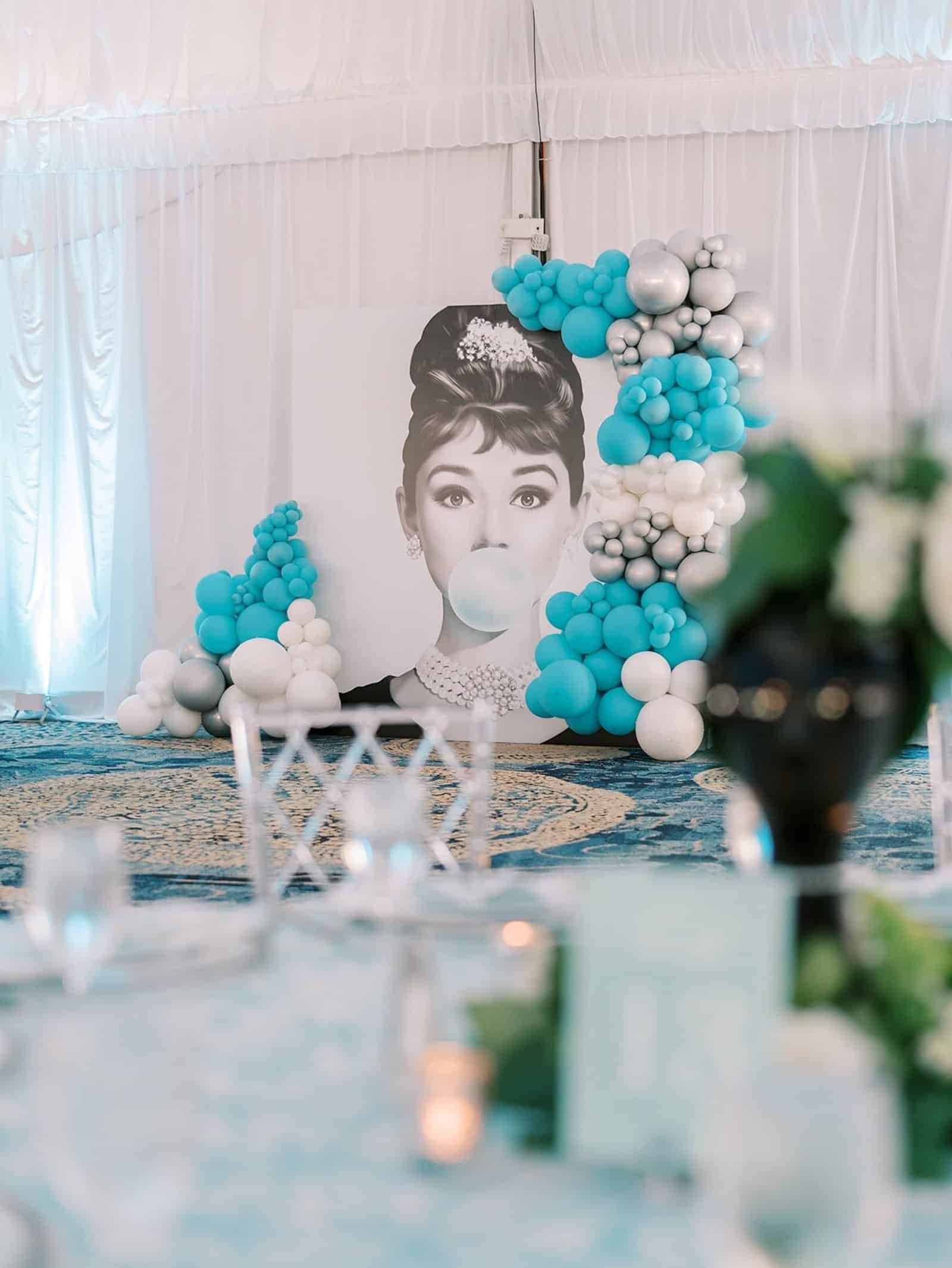 Audrey Hepburn bubblegum portrait backdrop framed by turquoise, silver and white balloon garlands, with clear chairs in the foreground