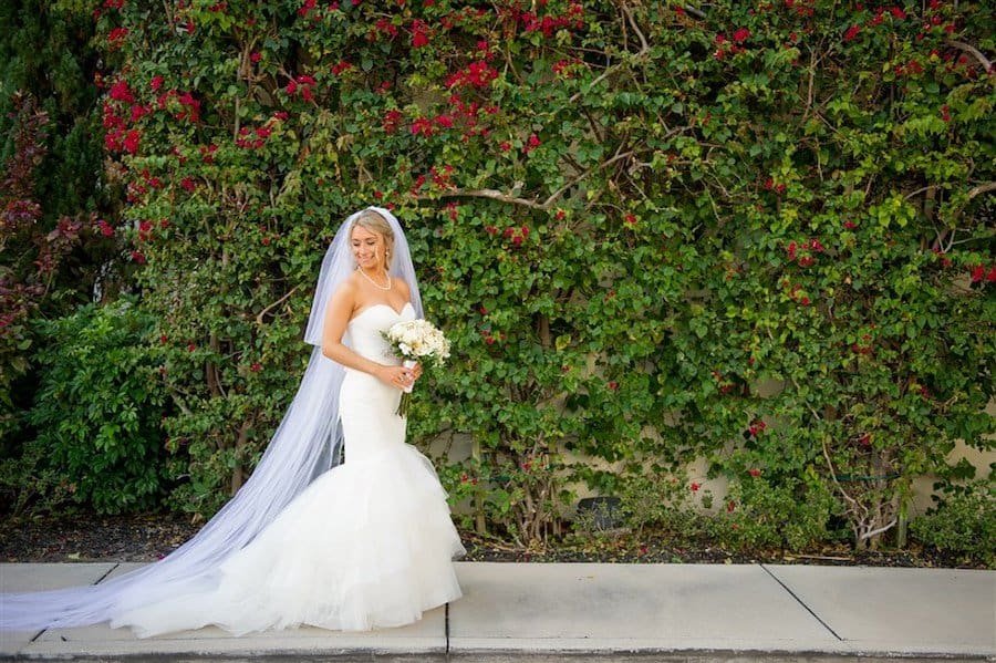 Bride in lace wedding gown holding vibrant fuchsia bouquet posing with groom in sharp black tuxedo on the golf course lawn