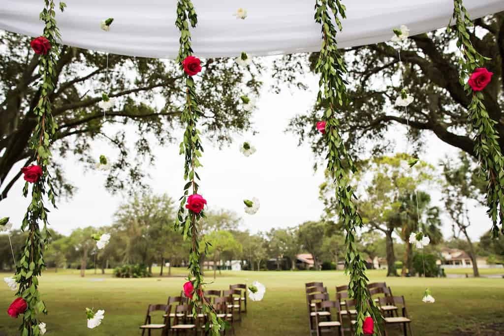 Outdoor Wedding Ceremony Arch and Wooden Folding Chairs at Countryside Country Club Outdoor golf course wedding ceremony arch with white sheer drapery, red rose garlands, and wooden folding chairs by Gabro Event Services at Countryside Country Club