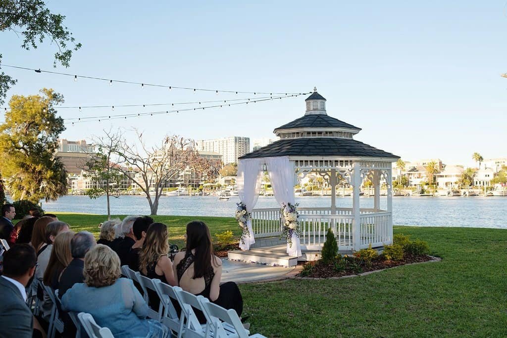 Davis Islands Garden Club outdoor ceremony setup with white folding chairs, waterfront gazebo with white draping, and overhead market bistro lighting by Gabro Event Services