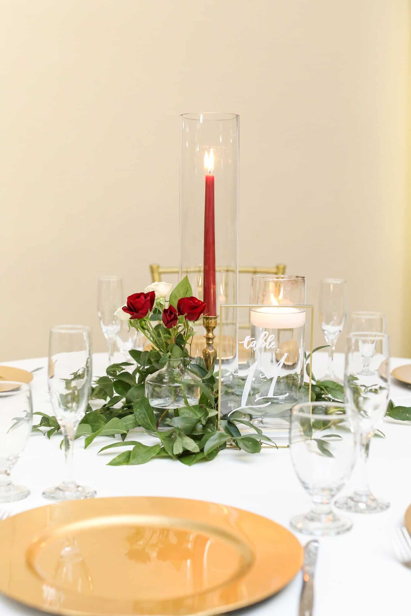 Close-up of elegant reception place setting featuring gold beaded charger plate, red rose floral compote, tall taper candlestick, and neutral tablecloths