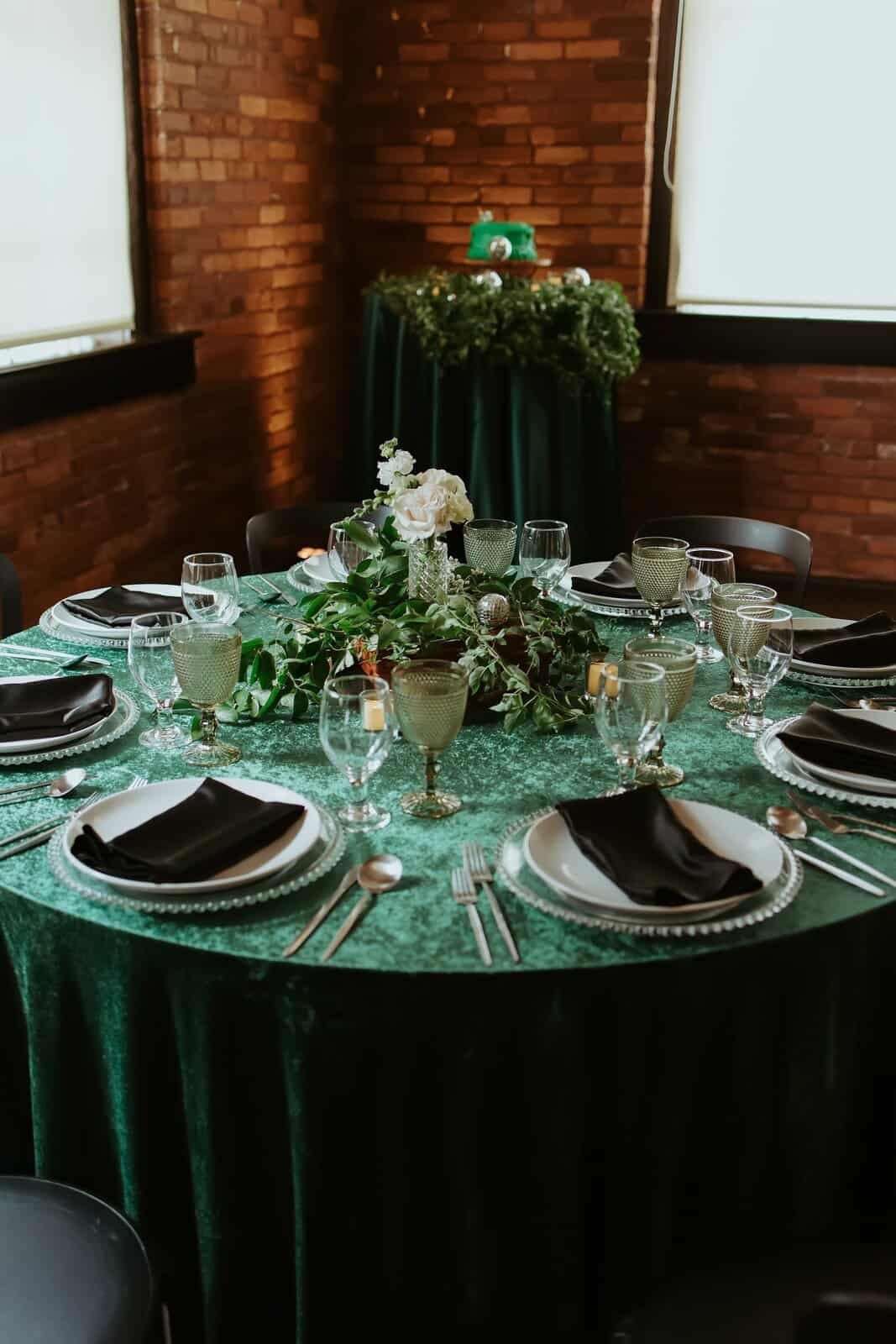Round table with emerald velvet linen, black napkins, green goblets and dark chargers in a brick room