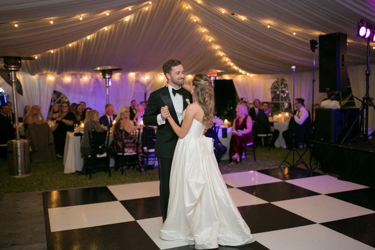 Bride and groom dancing on a black and white checkered dance floor under string lights and purple uplighting