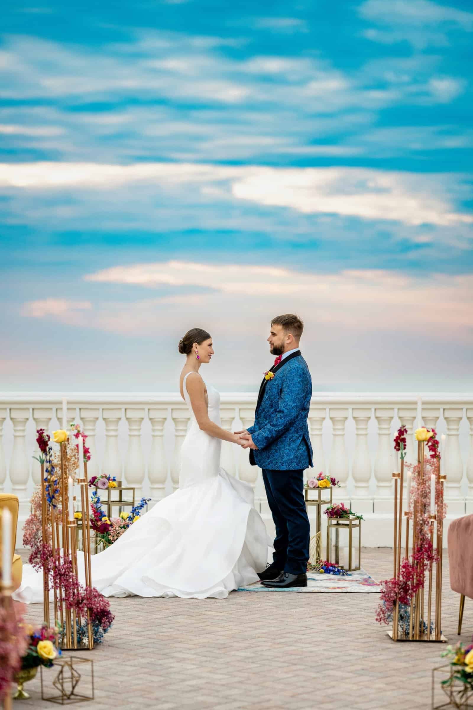 A bride in a form-fitting white gown standing before a sunset beach backdrop.