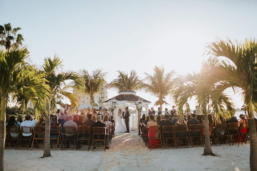 A bride and groom standing by the water during a sunset ceremony.