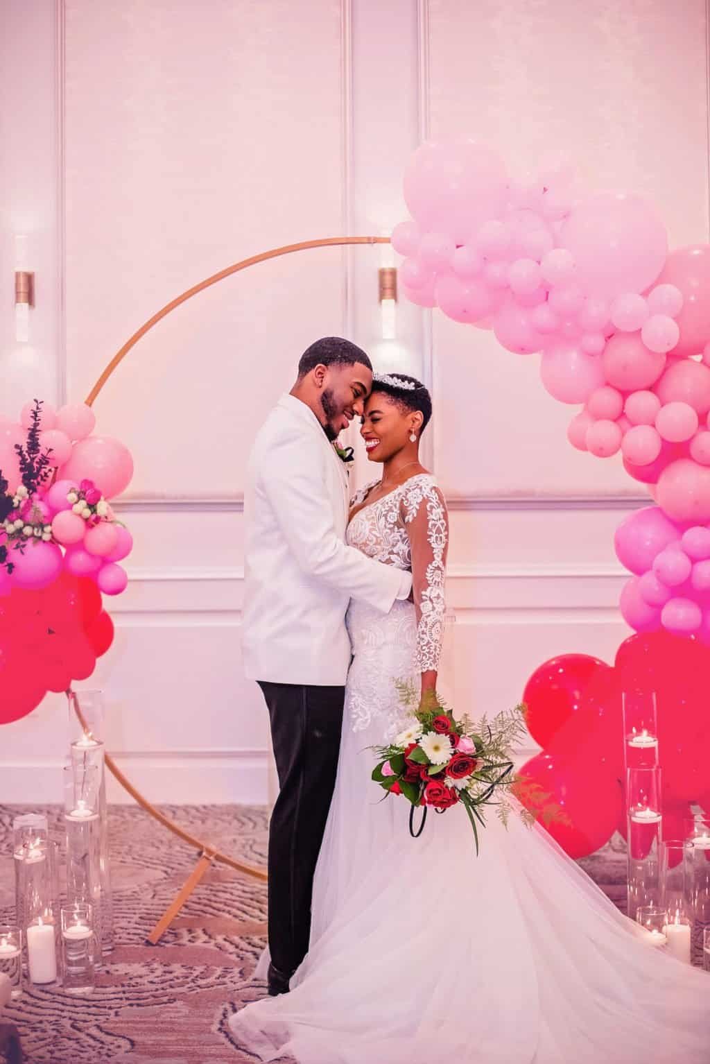 Bride in lace gown and groom in black tuxedo under modern pink and red ombre ceremony arch