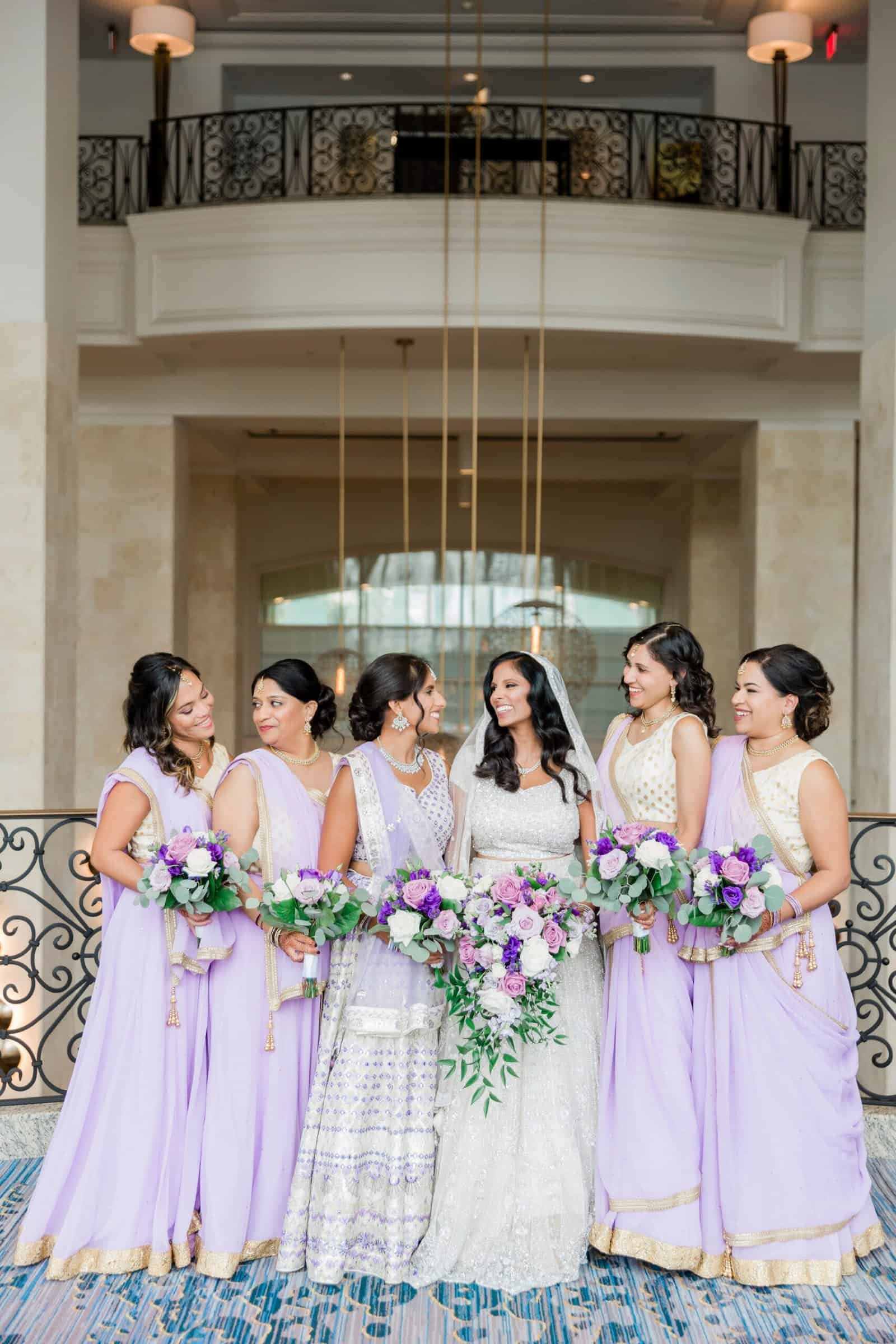 Bridesmaids in flowing lavender and violet silk sarees with floral bouquets