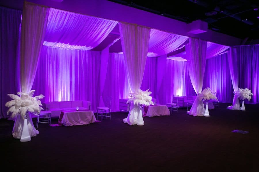 Plush modern wedding lounge area featuring curved white banquette seating, metallic throw pillows, and purple accent lighting