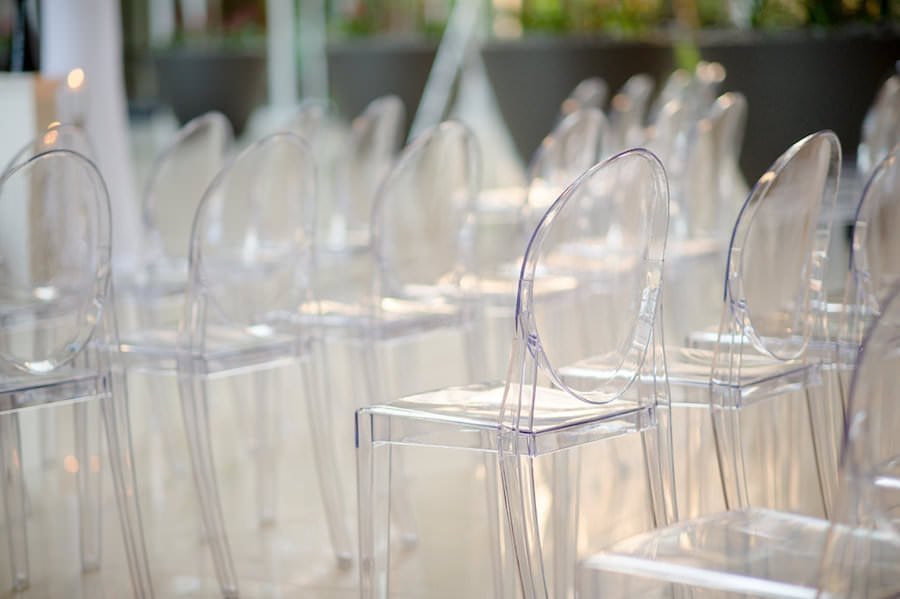 Sleek wedding ceremony aisle lined with transparent clear Ghost chairs, white floral pedestals, and draped columns