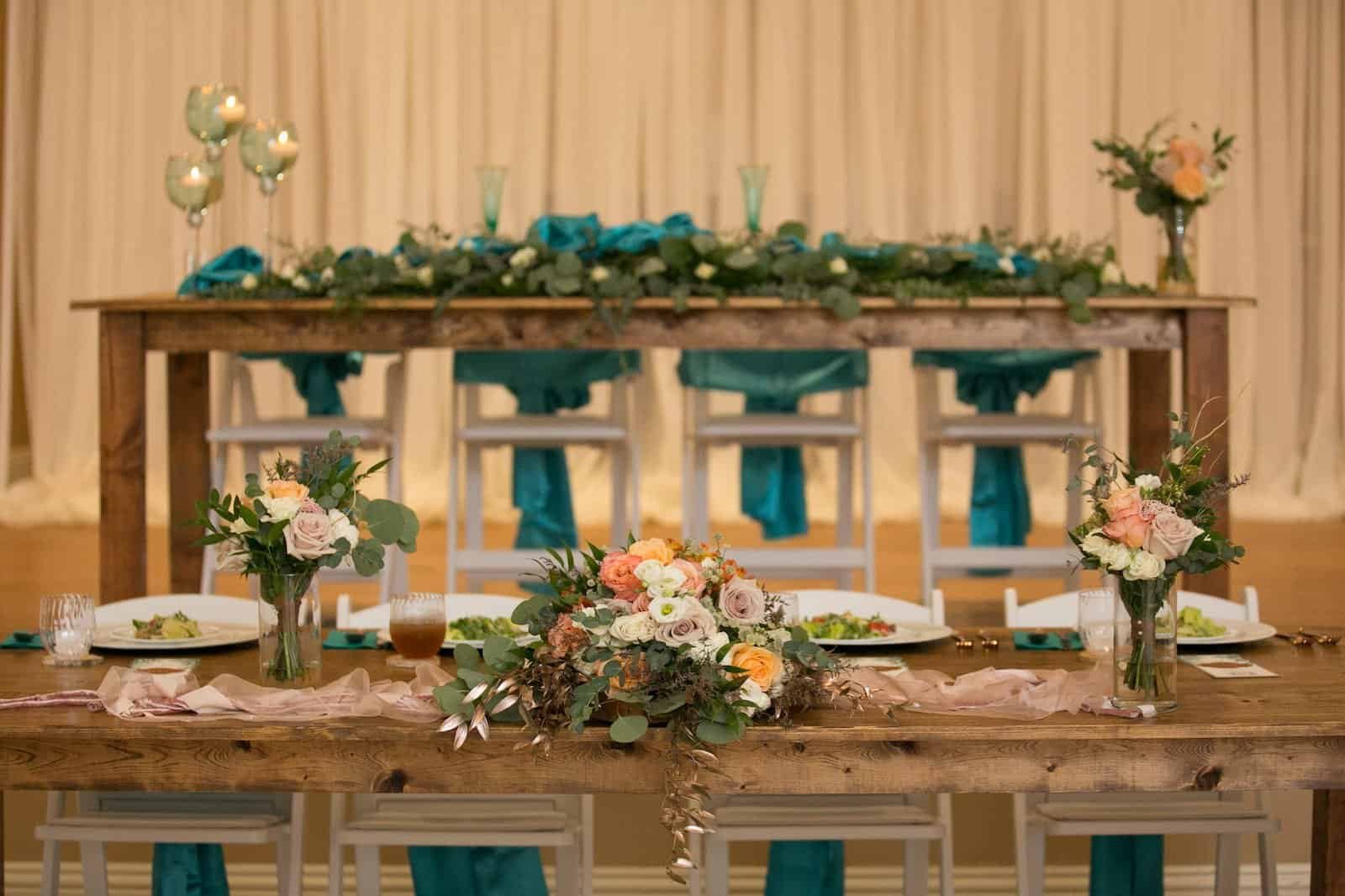 Rustic wedding reception at Shahnasarian Hall featuring long wooden farm tables decorated with teal chair sashes and custom linen rentals from Gabro Event Services
