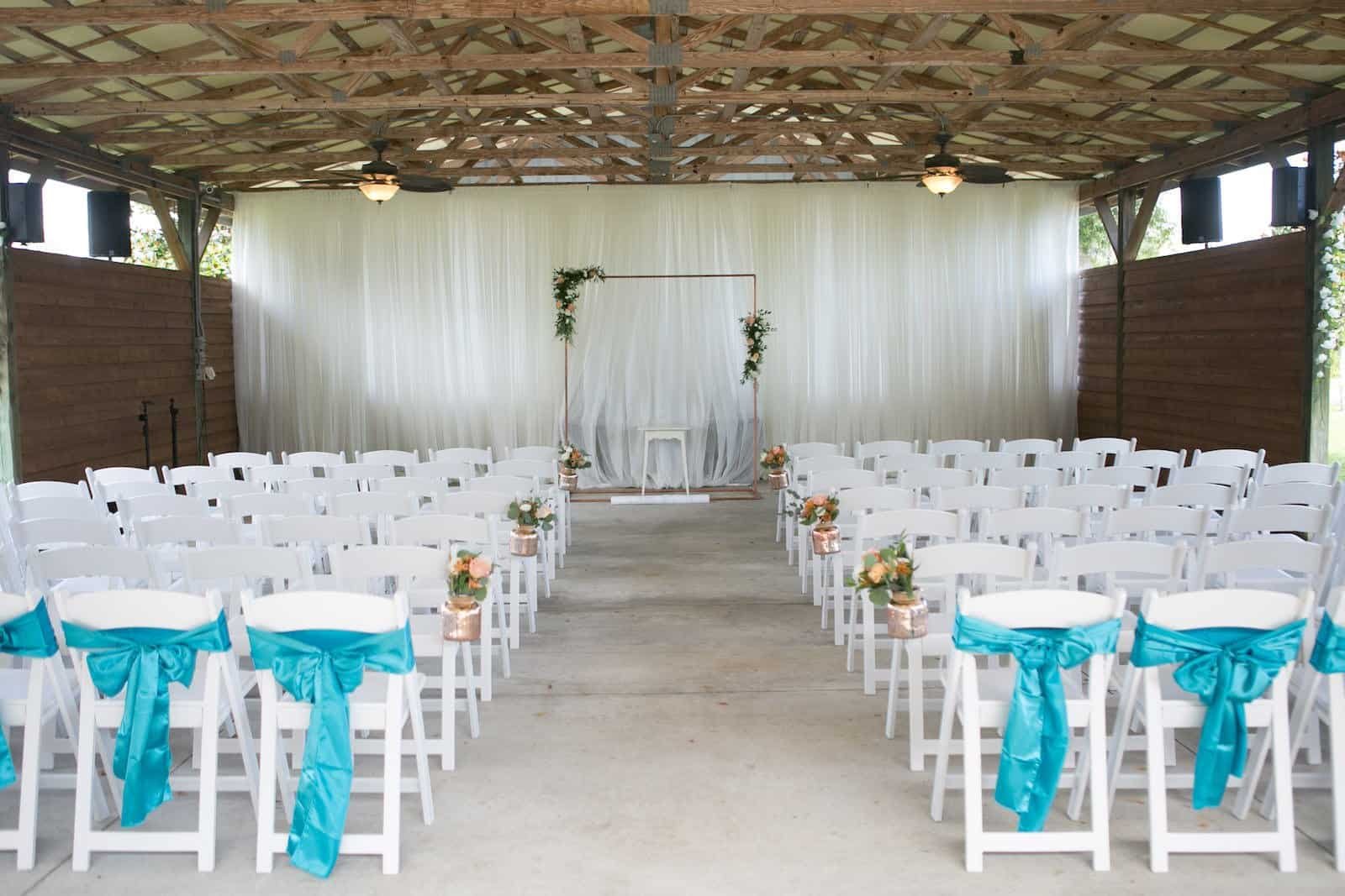 Pinellas Park wedding ceremony setup at Shahnasarian Hall featuring rectangular copper arch, gold mercury glass jars, and white folding chairs with teal ribbons