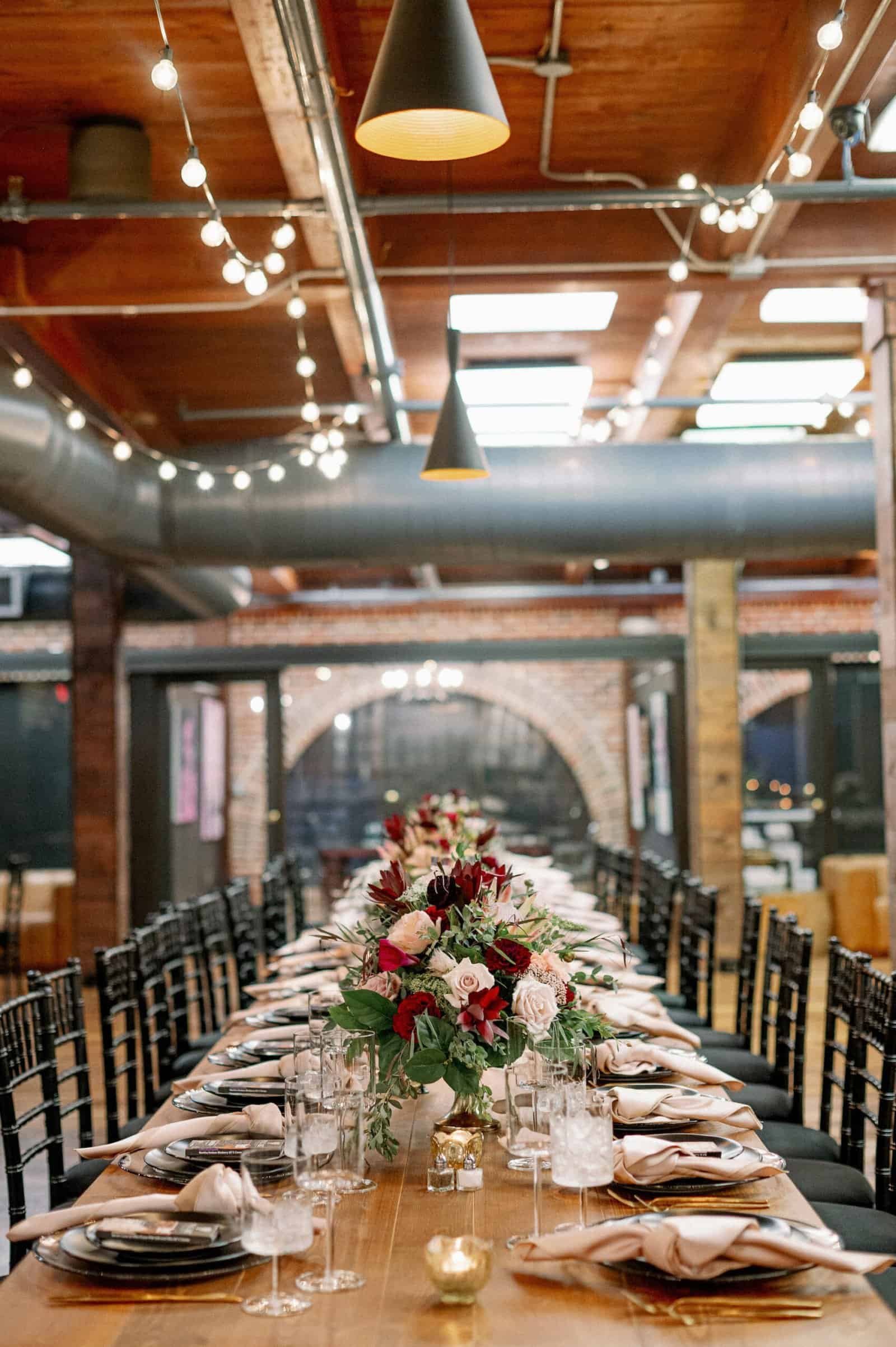 Long run of bare wood farm tables with black Chiavari chairs in the brick and timber room at Station House