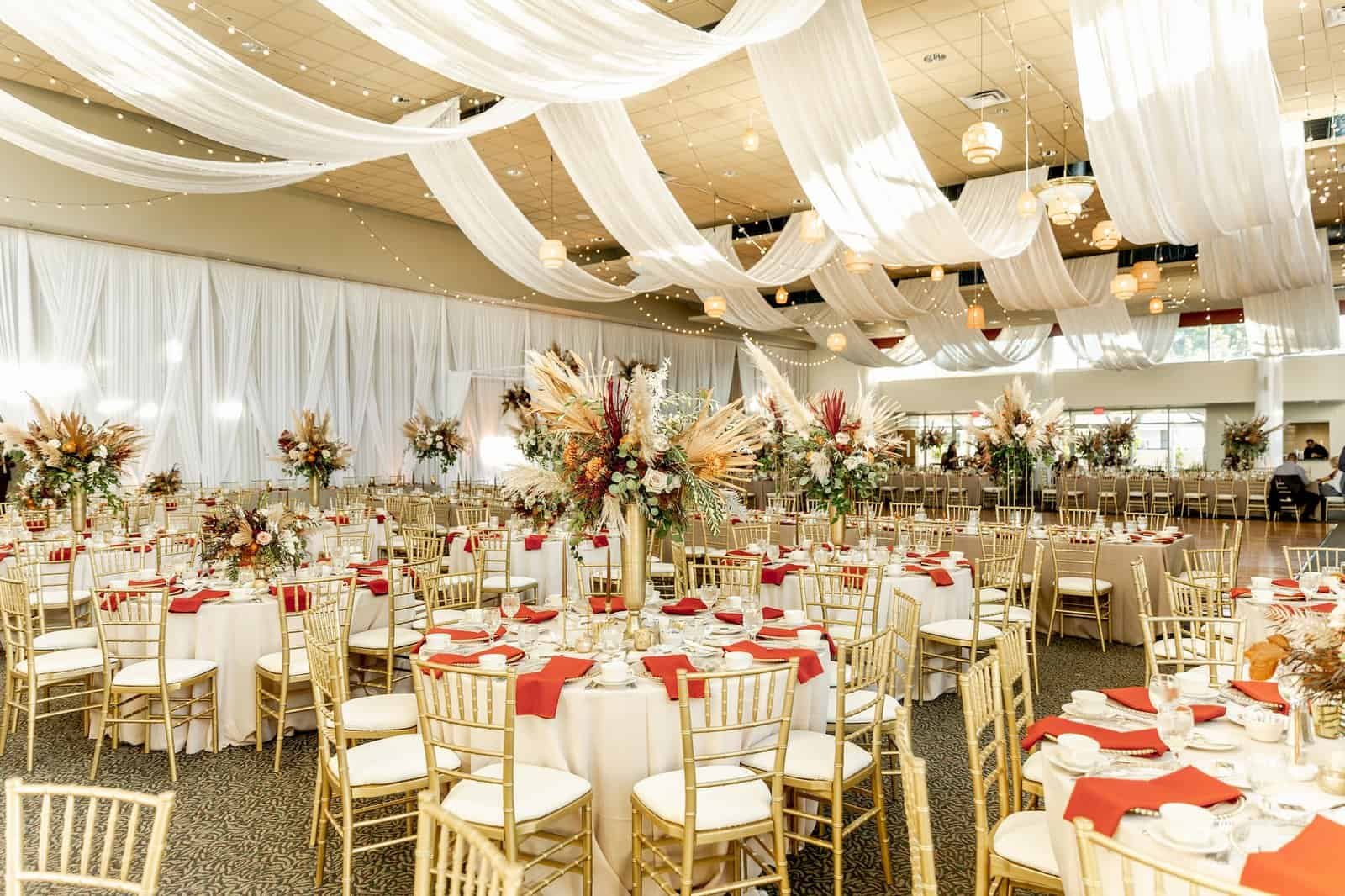 White ceiling drape radiating across the hall above round tables and gold Chiavari chairs