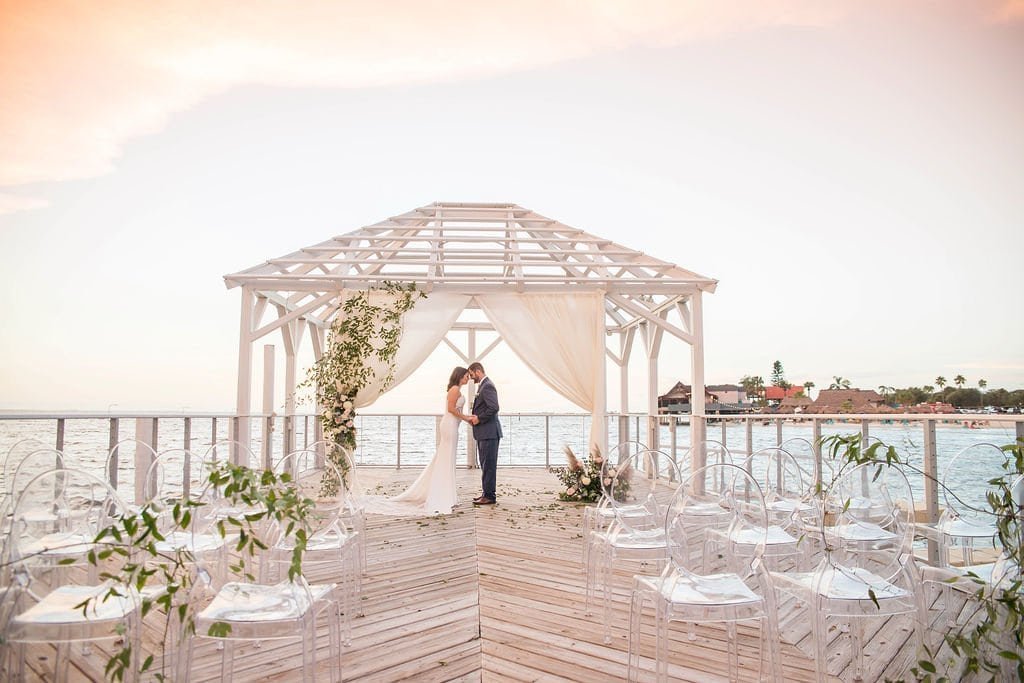 Bride and groom exchanging vows under white draped ceremony canopy with clear ghost chairs at The Godfrey