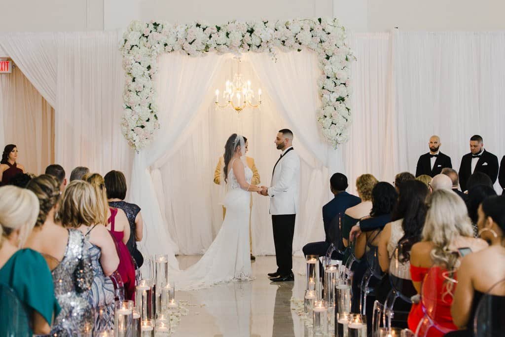 Bride and Groom Exchanging Wedding Vows Under White Draping Backdrop and Chandelier at The Vault
