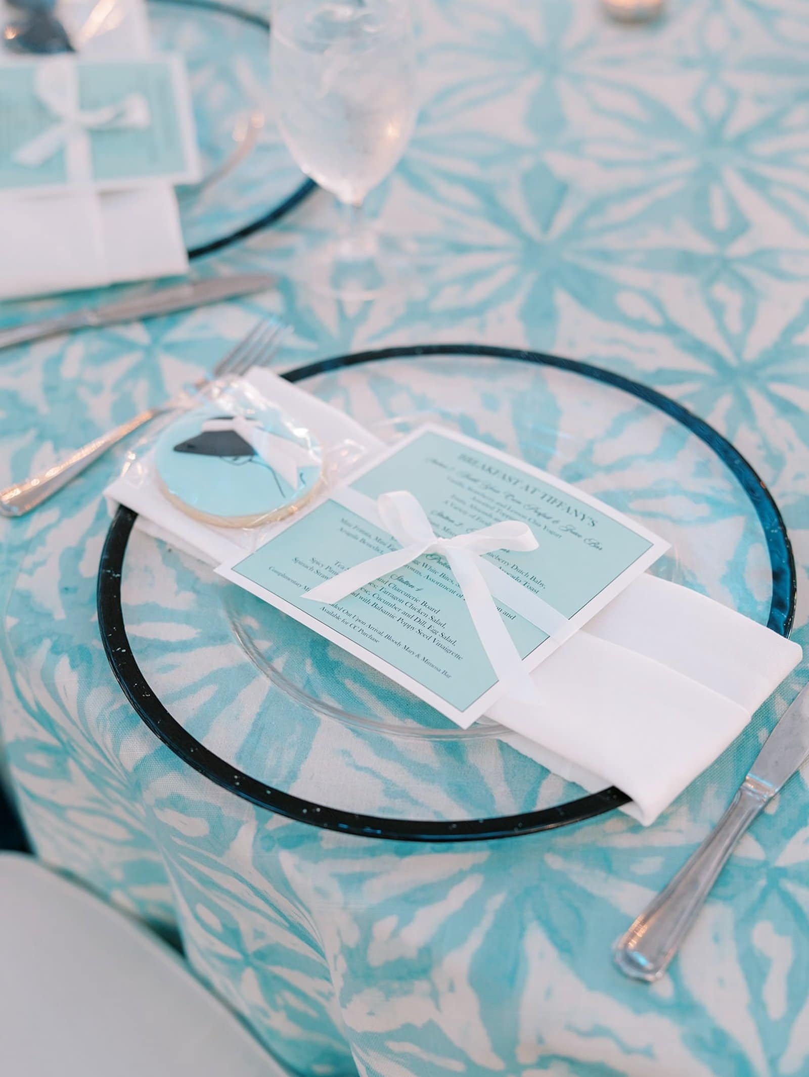 Clear glass charger with a black rim holding a white napkin, a Breakfast at Tiffany's menu card tied with white ribbon and a decorated cookie on an aqua patterned tablecloth