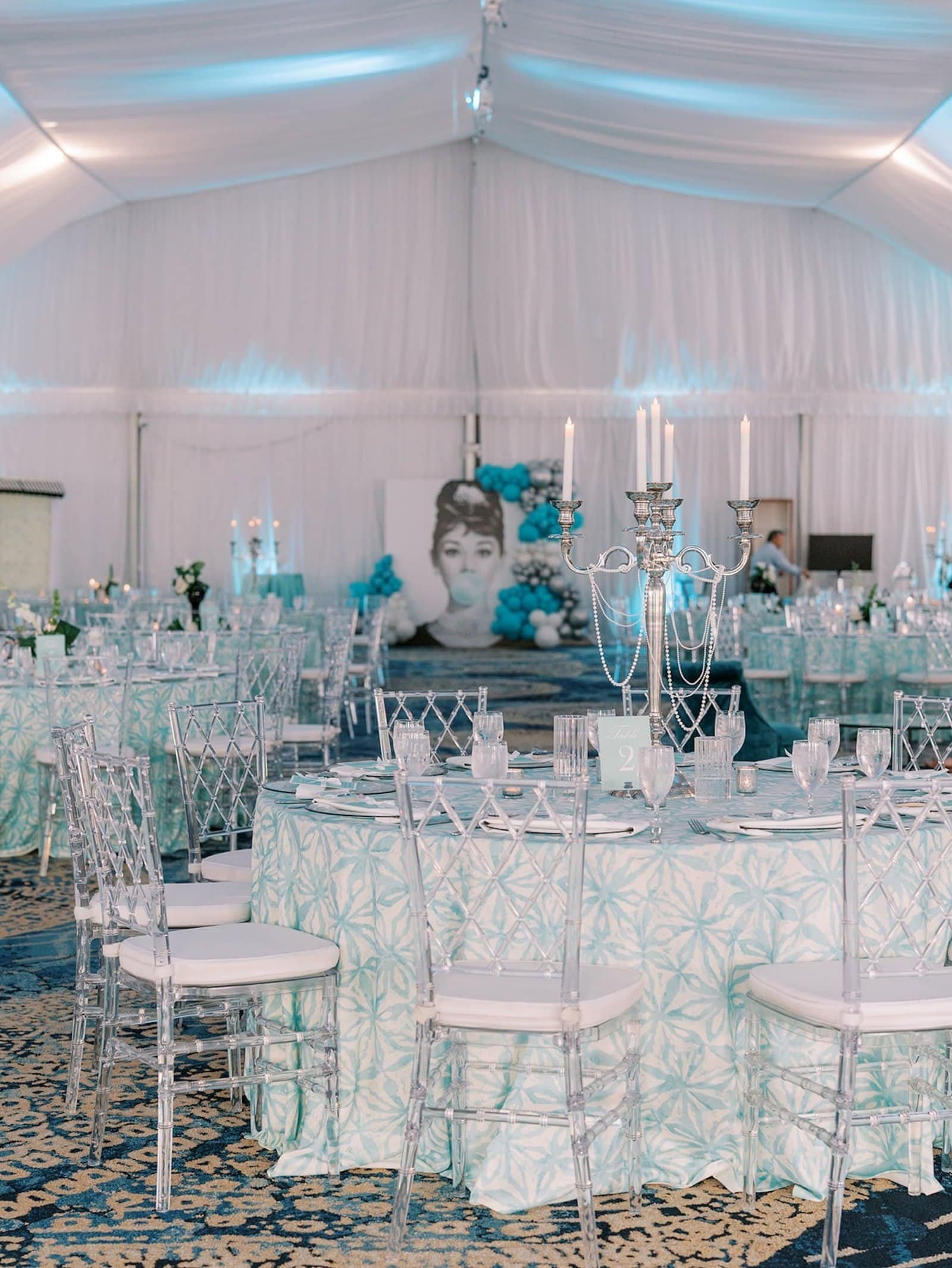 Round tables in aqua patterned linens with clear chairs and silver candelabras, an Audrey Hepburn backdrop beyond, under white ceiling drapery