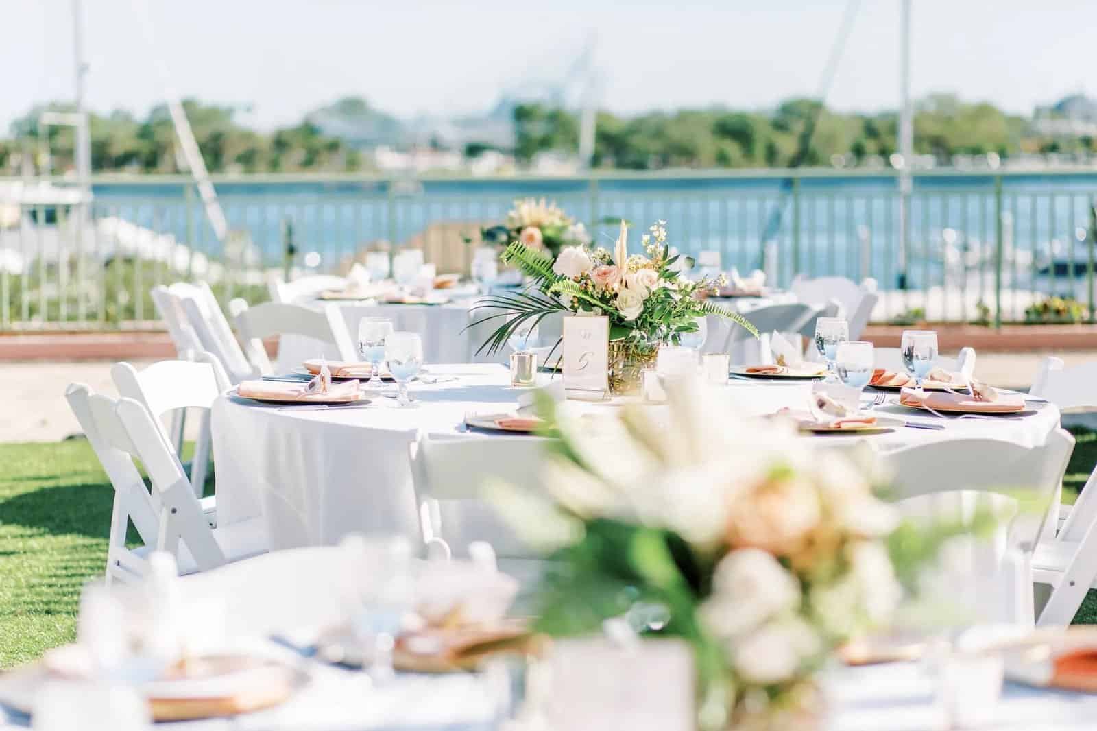 Round reception table with white linen, blush napkins, white garden chairs and low arrangements overlooking the marina